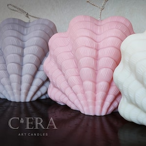 May include: Three shell-shaped candles in light purple, soft pink, and white, each featuring a textured ribbed surface and a top wick. The text C'ERA ART CANDLES is printed in white on the lower left side of the image, near the base of the candles.
