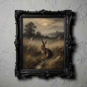 May include: A framed painting featuring a brown hare sitting in a field of tall grass under a full moon. The artwork is enclosed in an ornate, black, decorative frame with intricate floral carvings along the edges, set against a textured wall.
