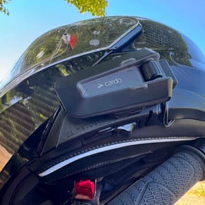 Op de afbeelding: Een zwarte motorhelm met een glanzende koolstofvezel afwerking en een helder vizier. Een zwart Cardo-communicatieapparaat is aan de zijkant van de helmschaal bevestigd. De helm is voorzien van een zwarte kinband en een zwarte handgreep met textuur.