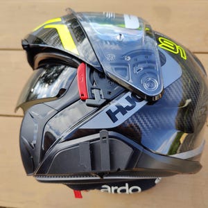 Könnte beinhalten: Ein schwarzer Motorradhelm aus Kohlefaser mit klarem Klappvisier, ausfahrbarer dunkler Sonnenblende und leuchtend gelben Akzenten. An der Seite befindet sich das HJC-Markenlogo in schwarzer Schrift sowie ein roter Entriegelungshebel und eine Halterung.
