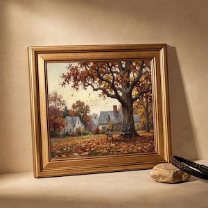May include: A framed landscape painting depicting an autumn scene with a large tree, fallen orange and brown leaves, a black wrought iron bench, and houses in the background. The artwork is enclosed in a thick, metallic gold-toned wooden frame.