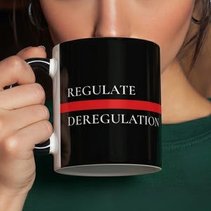 May include: Black and white ceramic mug with the words "REGULATE" above a red line and "DEREGULATION" below. The mug is held by a person. The background includes bookshelves and jars.