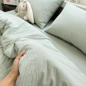 May include: A sage green and white gingham patterned duvet cover and matching pillowcases. The fabric features a small-scale checkered print. The bedding set is displayed on a bed with a plush white rabbit toy and a small black and white zebra figurine.
