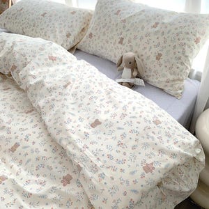 Könnte beinhalten: Cremefarbenes Bettwäsche-Set mit einem sich wiederholenden Muster aus kleinen Blumen und Teddybären. Das Set beinhaltet einen Bettbezug und zwei Kissenbezüge. Ein Stoffhase liegt auf dem Bett. Die Bettwäsche wirkt weich und einladend.