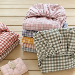 May include: A selection of folded bed sheets in a variety of colours and checkered patterns, including pink, blue, yellow, and green. The sheets are stacked on a light wooden surface. A small, pink quilted pouch with a decorative tag is in the foreground.