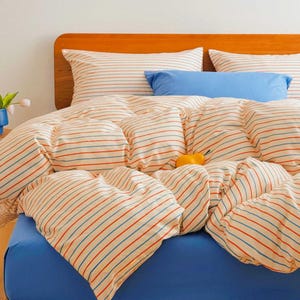 Op de afbeelding: Een bed met een houten hoofdbord en blauwe lakens. Het beddengoed heeft een crèmekleurig dekbed en kussenslopen met rode en blauwe horizontale strepen. Een blauw sierkussen ligt bovenop het dekbed.