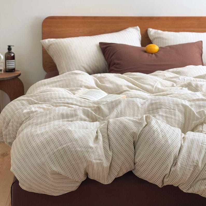 May include: A bed with a wooden headboard and a cream-colored striped duvet cover. Two white striped pillows and a brown pillow are on the bed. A lemon rests on the brown pillow. A book is open on the bed.