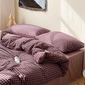 May include: A bed dressed with a duvet and two pillows, all in a pink and brown gingham check pattern. The bedding is arranged on a bed with a plain pink bed skirt. A decorative wall hanging and a wooden tree are in the background.
