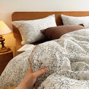 May include: A bed with a wooden headboard is covered with a floral patterned duvet and pillows. The duvet and pillowcases have a white background with blue and gray floral designs. A brown pillow and a lamp are also visible.