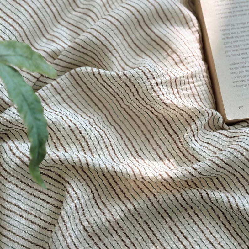 May include: Cream-colored fabric with thin, brown vertical stripes. The fabric is draped, creating soft folds and shadows. A green leaf and the corner of a book are visible in the image, suggesting a cozy, natural setting. The fabric appears to be lightweight.