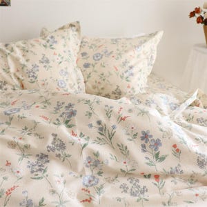 Peut inclure: Ensemble de literie crème avec un motif floral délicat. L'ensemble comprend deux taies d'oreiller et une housse de couette, ornées de fleurs bleues, violettes et rouges et de feuilles vertes. L'ensemble crée une ambiance douce et accueillante.