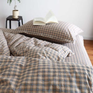 Pode incluir: Conjunto de roupa de cama com padrão xadrez bege e azul. O conjunto inclui uma almofada, uma capa de edredão e um lençol com elástico. Um livro aberto repousa sobre a almofada. A roupa de cama é feita de um material macio e confortável.