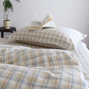May include: A bed set with a plaid pattern in beige, blue, and brown. The set includes a pillow and a sheet. An open book rests on the pillow. The bedding is made of a soft, comfortable material.
