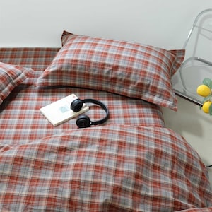Pode incluir: Um conjunto de roupa de cama com padrão xadrez em tons de laranja queimado, cinzento e branco. O conjunto inclui uma fronha retangular e uma capa de edredão a condizer. Um livro branco e auscultadores pretos estão sobre a cama.
