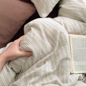 May include: Close-up of a cream-colored duvet cover with thin, brown vertical stripes. The bedding is on a bed with a brown pillow and a book. The duvet cover is being held by a hand.