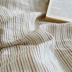 May include: Close-up of a cream-colored fabric with vertical brown and green stripes. The fabric is draped and folded, creating a textured appearance. A book with yellowed pages is visible in the upper right corner.