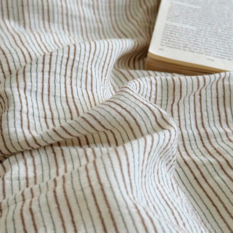 May include: Close-up of a cream-colored fabric with vertical brown and green stripes. The fabric is draped and folded, creating a textured appearance. A book with yellowed pages is visible in the upper right corner.