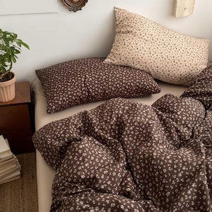 Peut inclure: Un lit avec une housse de couette à motifs floraux bruns et deux taies d'oreiller assorties. Une taie d'oreiller est marron et l'autre beige. La literie présente un petit motif floral blanc. Le lit est fait avec un drap de couleur neutre.