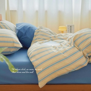 May include: A bedding set featuring a solid blue fitted sheet, a solid blue pillowcase, and a duvet cover with a cream and blue striped pattern. The text "No alarm clock, no noise, no work, Slow down and live well." is printed on the blue sheet.