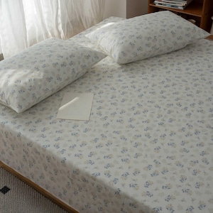 Könnte beinhalten: Ein Bett mit weißem Bettlaken und zwei passenden Kissenbezügen, alle mit kleinen blauen und beigen Blumenmustern. Eine weiße Karte liegt auf dem Laken. Schlafzimmer-Atmosphäre.