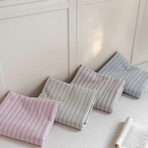 May include: Four folded striped pillowcases in pastel colors: pink, green, lavender, and blue. Each pillowcase has thin, dark stripes. The pillowcases are arranged in a row, leaning against a white wall.