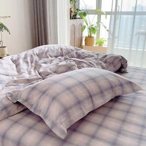 May include: Bedding set featuring a plaid pattern in pink, grey, and white. The image shows a pillow and a duvet cover, neatly arranged on the bed. The room has a window with curtains and plants in the background.