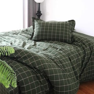 May include: A green and white checkered bedding set, including a pillow and comforter, is displayed on a bed. The fabric has a grid pattern. The scene is lit by natural light, suggesting a bedroom setting. A fern is visible in the lower left corner.