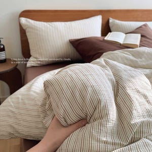 May include: A bed with a wooden headboard, featuring striped bedding in cream and brown. The pillows are cream and brown. The text on the image reads: "Lying down is like being completely enveloped by clouds, in a state of total relaxation."
