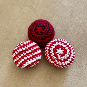 May include: Three round, crocheted spheres. One sphere features a dark red and black spiral pattern. The other two spheres display a vibrant red and white striped pattern, with one showing a circular center design and the other a diagonal stripe.