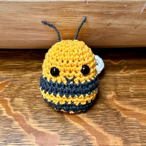 May include: A handmade crochet bee with yellow and dark gray stripes. The bee has black eyes and two black antennae. A small white wing is visible on the side. The bee is sitting on a wooden surface.