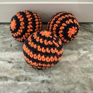May include: Three spherical objects crafted from yarn, each featuring a repeating pattern of alternating black and bright orange stripes. The crochet texture is visible across the surface of these round items, which are arranged in a triangular formation.
