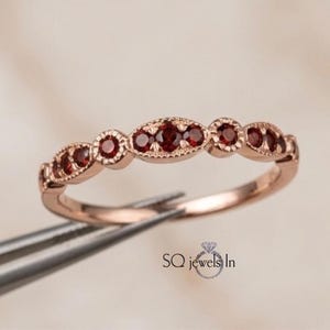 Op de afbeelding: Rosegouden ring met afwisselende ronde en ovale zettingen, elk met een kleine, dieprode edelsteen. Het ontwerp van de ring wordt benadrukt tegen een neutrale achtergrond, waarbij de kleur en details van de edelstenen worden benadrukt.