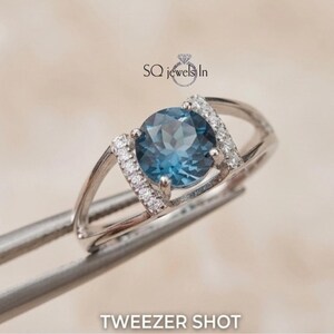 Può includere: Anello d'argento con una grande gemma blu rotonda al centro, affiancata da pietre più piccole e trasparenti. L'anello è tenuto da una pinzetta. Il testo "SQ jewels In" e "TWEZER SHOT" è visibile.