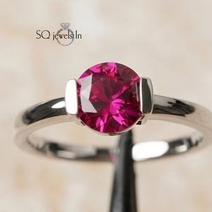 Könnte beinhalten: Ein silberfarbener Ring mit einem runden, leuchtend fuchsia-pinkfarbenen Edelstein in einer Spannfassung. Die Ringschiene ist glatt und poliert. Der Text SQ jewels In ist oben links im Bild zu sehen, begleitet von einem Diamantsymbol.