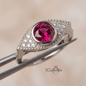 Op de afbeelding: Een zilverkleurige ring met een centrale ronde, felroze edelsteen in een zetting, omringd door een marquisevormig frame bezet met kleine heldere steentjes. De tekst SQ jewels In staat in het zwart onderaan de afbeelding.