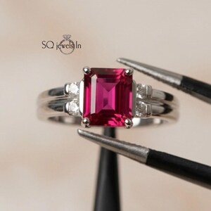 May include: A silver-toned ring featuring a central emerald-cut vibrant pink gemstone flanked by two small round clear stones on each side. The metal band has a polished finish. The text SQ jewels In is visible in the upper left corner.