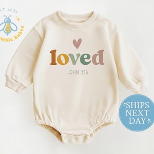 May include: Cream-colored baby onesie with long sleeves. The word "loved" is printed in colorful letters, with a heart above it. The text "JOHN 3:16" is below. A logo with a bee and the words "Us Bumble Babee" is in the upper left corner.