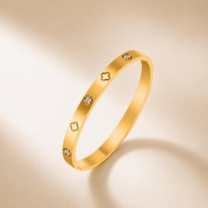 May include: A gold-toned bangle bracelet featuring a repeating pattern of alternating circular cutouts and small, clear, round-cut stones set flush into the metal band. The bracelet has a smooth, polished finish and a thin, circular profile.