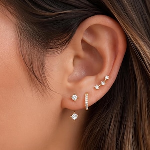May include: A set of three gold-toned earrings featuring clear stones. One earring is a small hoop with pavé stones, one is a single square-cut stud with a dangling square stone, and one is a curved bar stud with three small round stones.