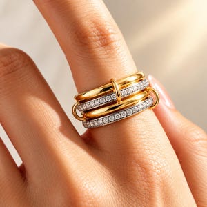 May include: A multi-layered ring featuring alternating bands of polished gold-tone metal and silver-tone metal encrusted with small clear stones. The bands are connected by a small vertical gold-tone metal loop, creating a stacked appearance.