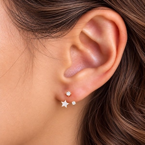 May include: A gold-toned earring set featuring three distinct elements: a small round stud, a larger round stud, and a star-shaped stud, all encrusted with clear, sparkling stones. The jewellery is positioned on the earlobe, showcasing a delicate design.