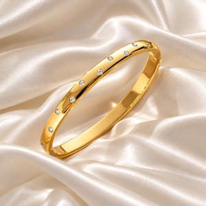 以下が含まれることがあります： 光沢のあるゴールドトーンのヒンジ付きバングルブレスレット。バンドの上半分に沿って小さな丸い透明な石が並んでいます。ブレスレットは滑らかで反射する表面を持ち、側面に留め金が見えます。
