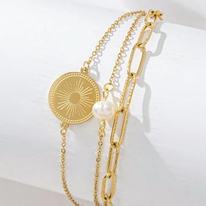 May include: Gold-coloured layered bracelet. It features a sunburst medallion, a delicate chain with a pearl, and a chunky link chain. The bracelet is displayed on a white surface, highlighting its intricate design and warm colour.