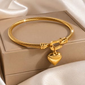 May include: A gold-toned cable-style bangle bracelet featuring a polished metal heart charm suspended from a U-shaped clasp mechanism. The bracelet has a twisted rope texture throughout the band, creating a continuous circular design with a metallic finish.