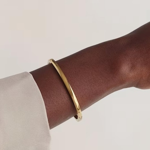 May include: A gold bangle bracelet. The bracelet has a simple, curved design and is worn on a person's arm. The arm is dark-skinned, and the person is wearing a light-coloured shirt. The background is a solid, neutral colour.