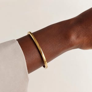 May include: A polished gold-tone bangle bracelet with a smooth, rounded surface and a visible hinged clasp mechanism. The bracelet features a simple, continuous circular design that rests securely around the wrist, showcasing a reflective metallic finish.