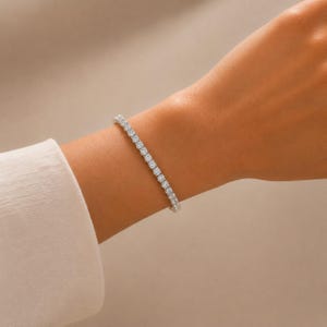 May include: A silver-toned tennis bracelet featuring a continuous row of round, clear, faceted stones set in a metallic frame. The bracelet encircles the wrist, showcasing a uniform design with sparkling, reflective surfaces throughout the piece.