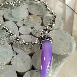 Op de afbeelding: Een zilverkleurige ketting met een grote, gladde, paarse hoornvormige hanger. De bovenkant van de hanger is afgewerkt met een donkere, getextureerde metalen band die is ingelegd met kleine, glinsterende, heldere steentjes die het licht vangen.