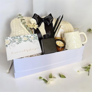 May include: A white gift box filled with a cream-colored blanket, a black bow, a mug, a candle, a reed diffuser, and a greeting card that reads "Especially for you". The box contains dried flowers and decorative elements.