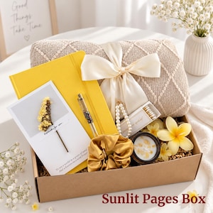 May include: A gift box containing a yellow notebook, a gold pen, a beige textured blanket with a cream ribbon, a pearl bracelet, a small candle, a gold scrunchie, a card with a dried flower, and a yellow flower. The text reads Sunlit Pages Box.